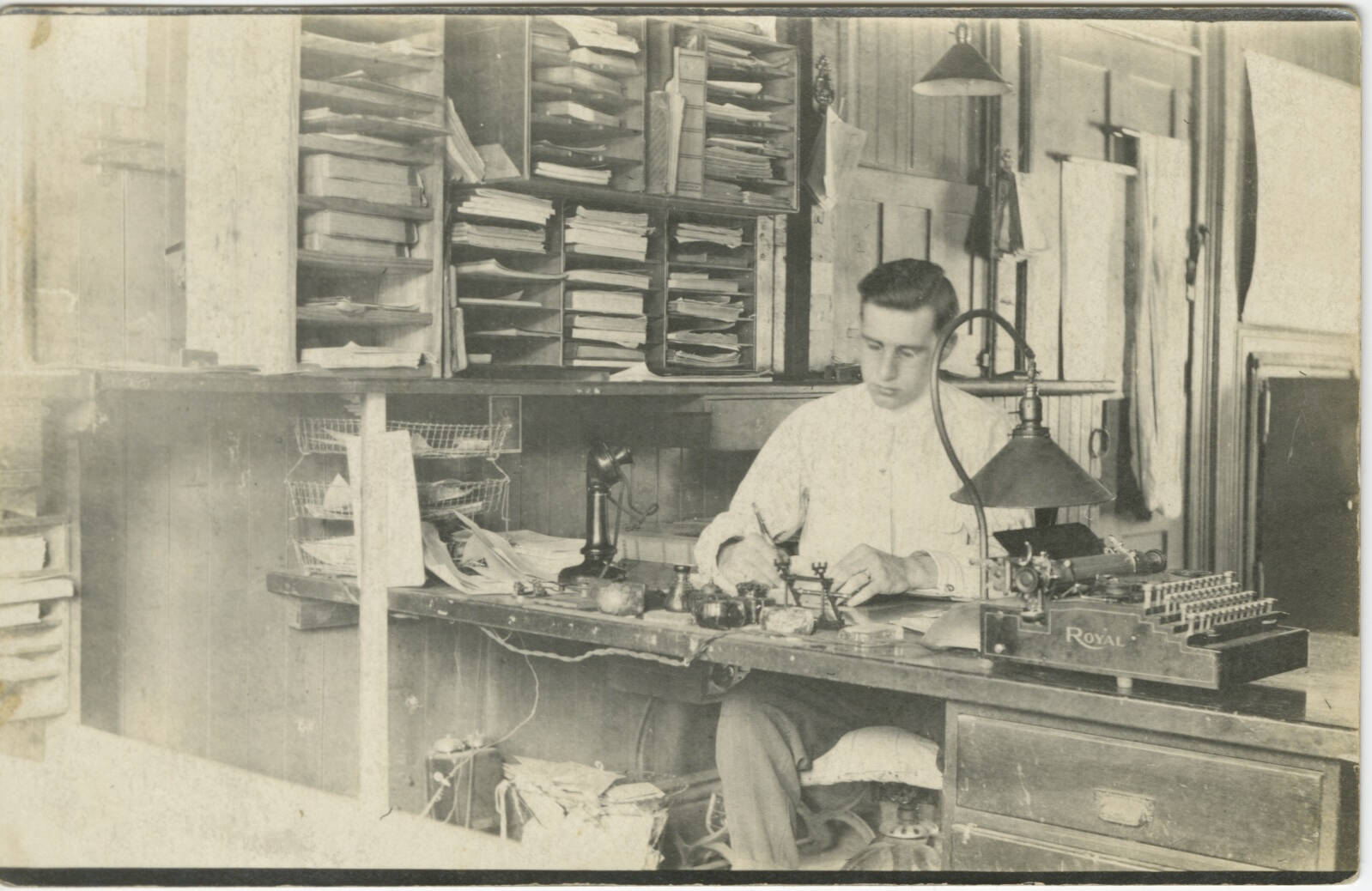Black and white real photo postcard of a man sitting at a desk in an office space.